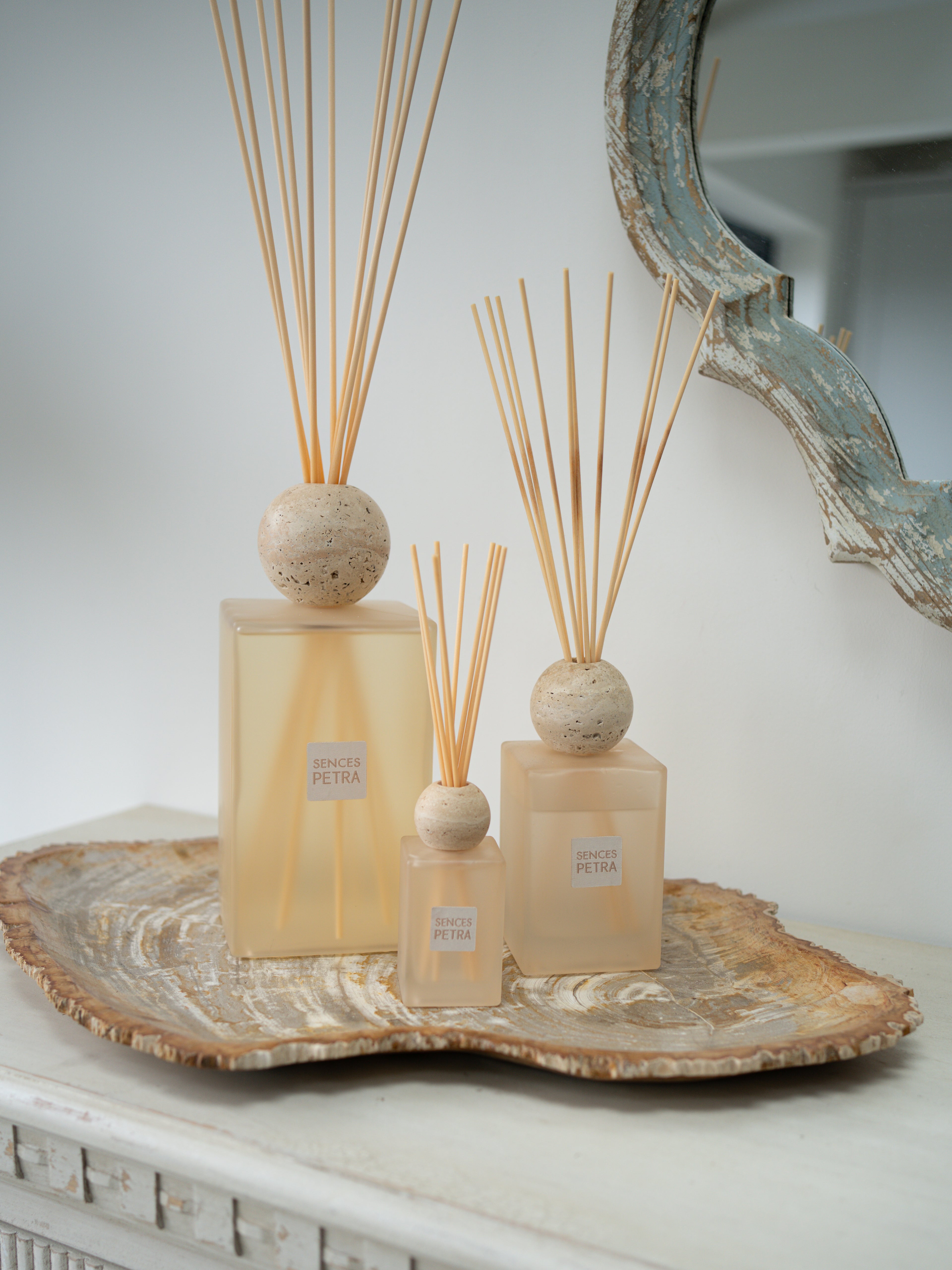 Three petra reed diffusers in frosted beige glass with natural wood reeds, displayed on a rustic silver tray against a light gray wall.