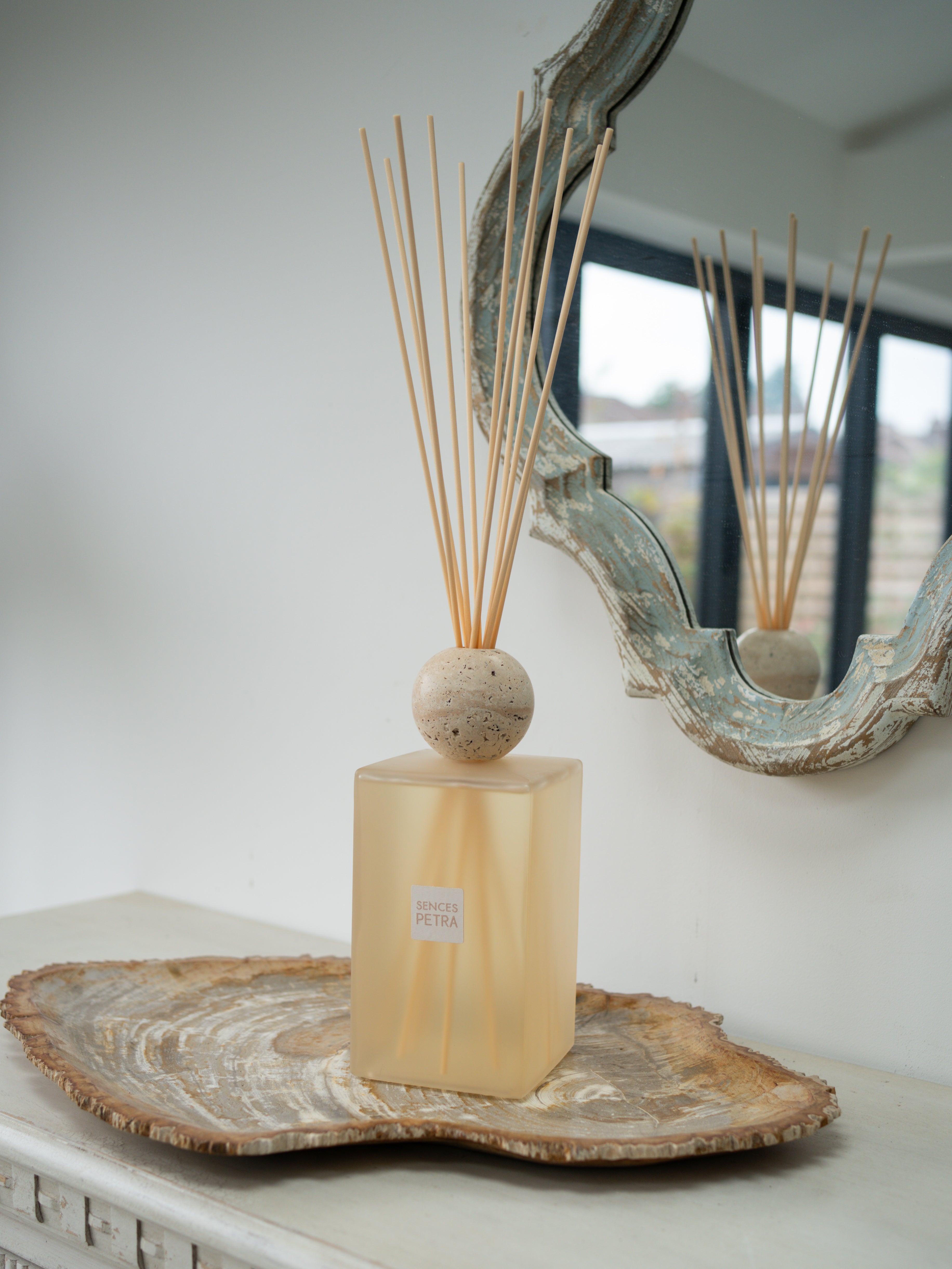 A reed diffuser with a translucent pale blush  rectangular bottle, a round beige travertine stone top, and natural reed sticks, displayed on a fossilled wood slice tray in front of a mirror.