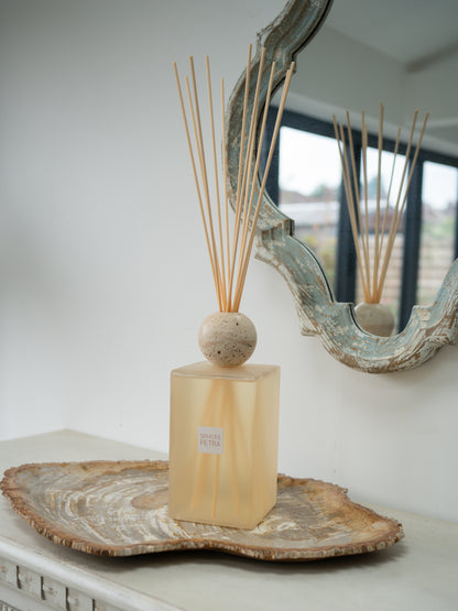 A reed diffuser with a translucent pale blush  rectangular bottle, a round beige travertine stone top, and natural reed sticks, displayed on a fossilled wood slice tray in front of a mirror.
