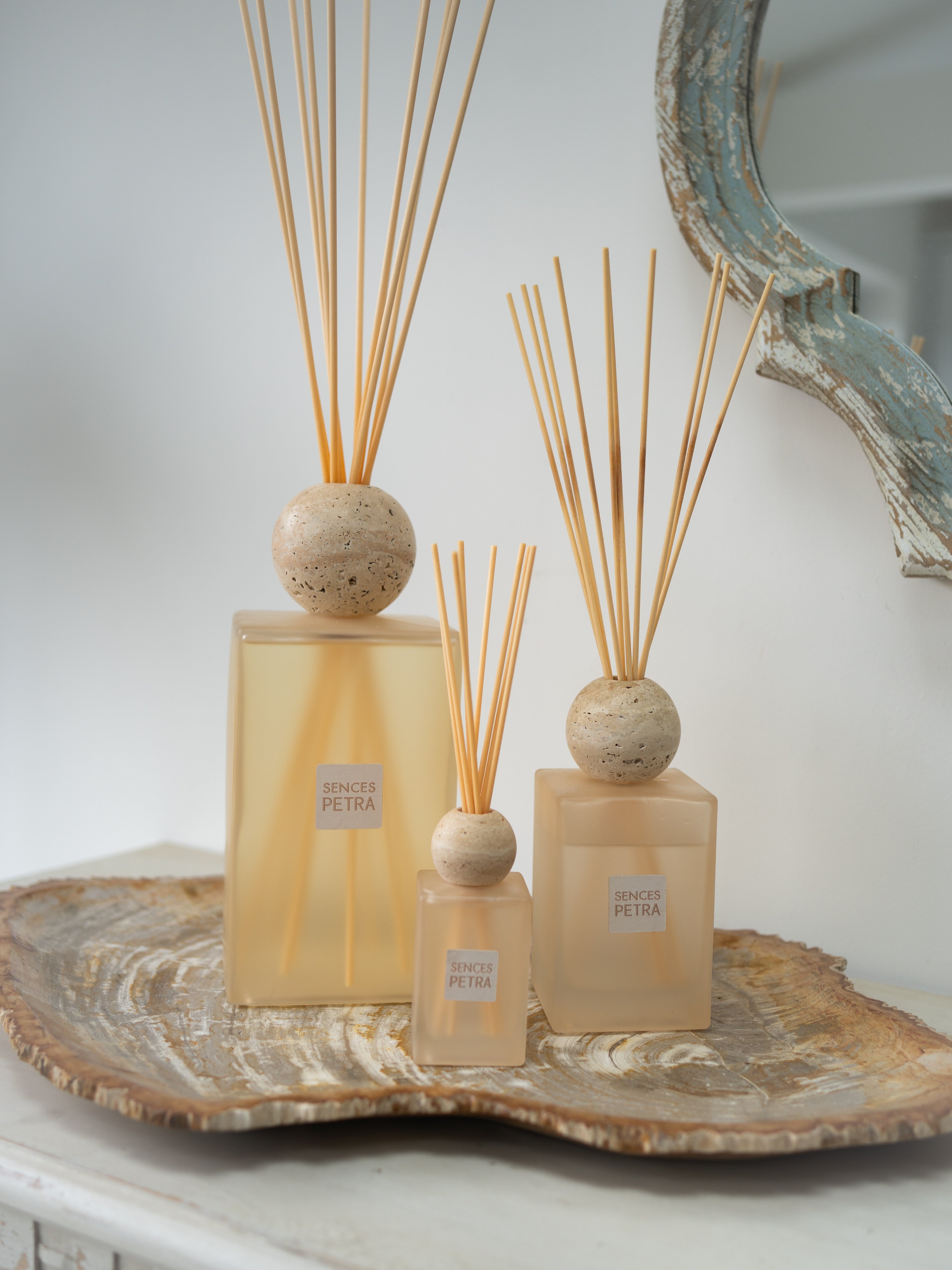 Three petra sences reed diffusers in varying sizes with beige square glass bottles, natural stone travertine tops, and light wood reeds, arranged on a decorative agate tray against a soft white background.