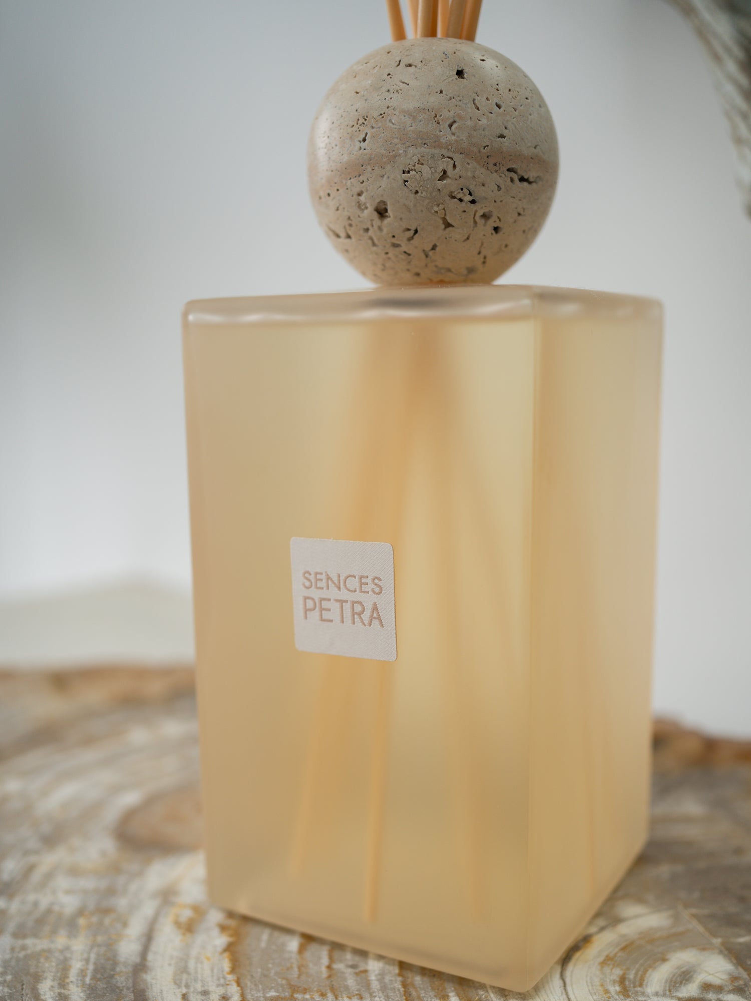 A Sences Petra reed diffuser with a translucent frosted beige glass bottle, natural fossilled wood diffuser sticks, and a round stone top, resting on a rustic wooden surface.