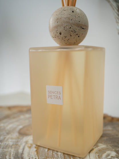 A Sences Petra reed diffuser with a translucent frosted beige glass bottle, natural fossilled wood diffuser sticks, and a round stone top, resting on a rustic wooden surface.