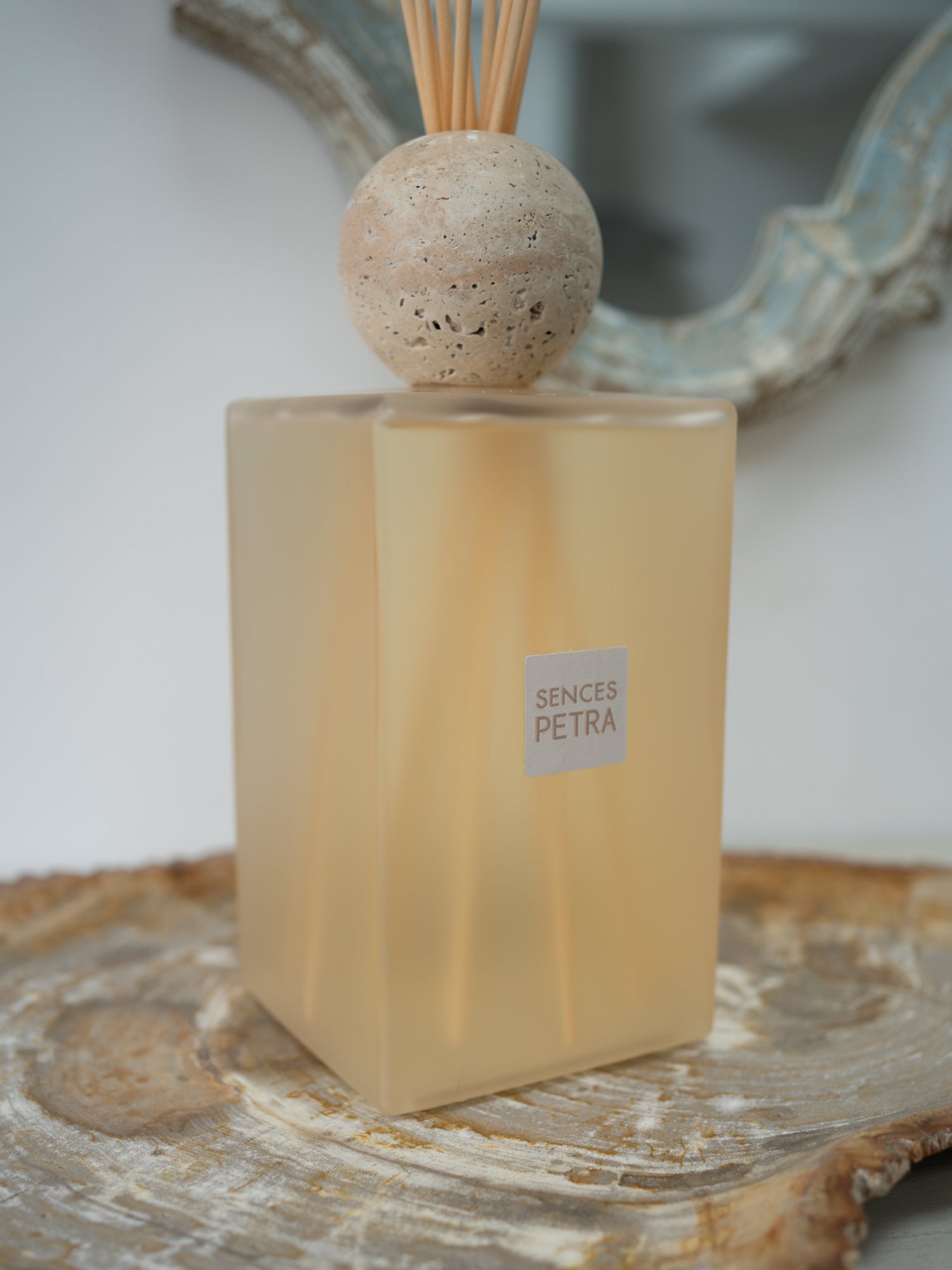 A reed diffuser bottle with a frosted warm beige glass body, a round natural travertine stone top, and wooden reeds, placed on a rustic wood slice with a blurred mirror in the background. The label reads &
