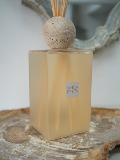 A reed diffuser bottle with a frosted warm beige glass body, a round natural travertine stone top, and wooden reeds, placed on a rustic wood slice with a blurred mirror in the background. The label reads &