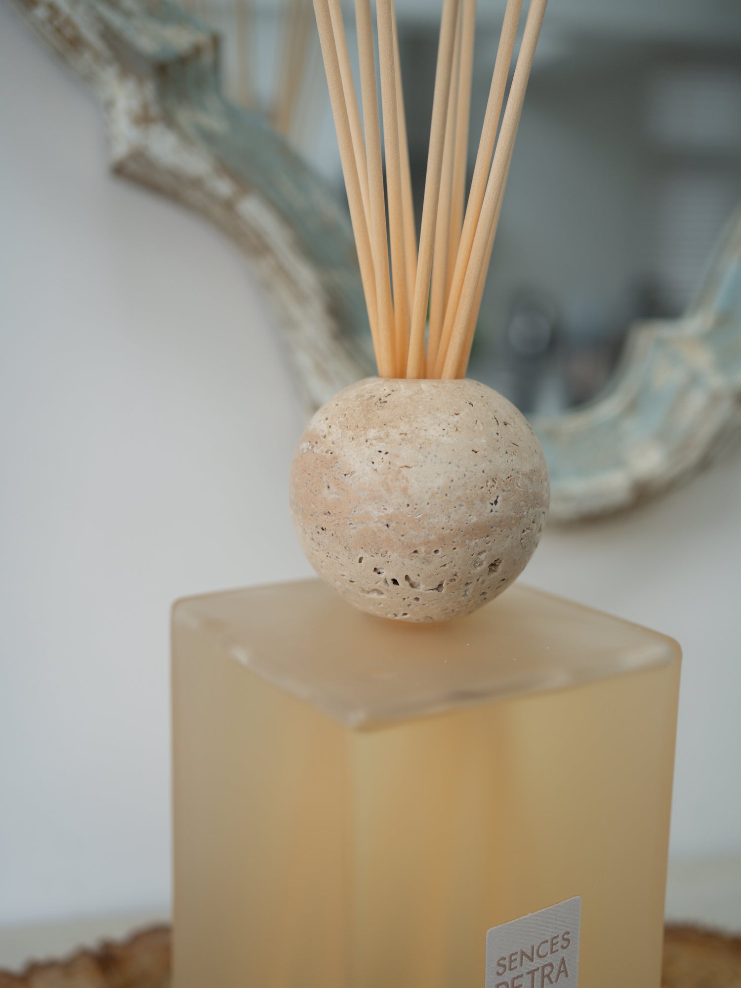 A reed diffuser with a round beige travertine stone sphere on a frosted amber glass base, with natural reed sticks inserted, photographed indoors against a softly blurred background.