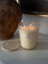 A lit candle in a frosted glass jar with three flames visible, placed on a light surface beside a round stone coaster. The candle label reads &