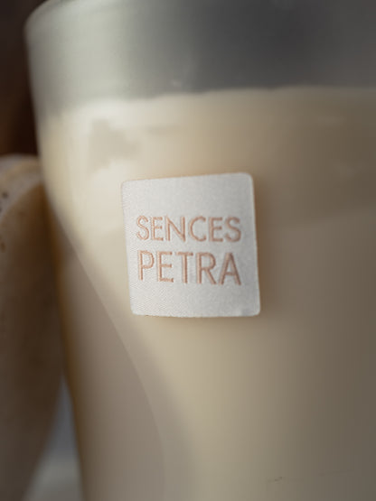 A close-up of a candle in a frosted glass container with a cream-colored wax and a white square label that reads &
