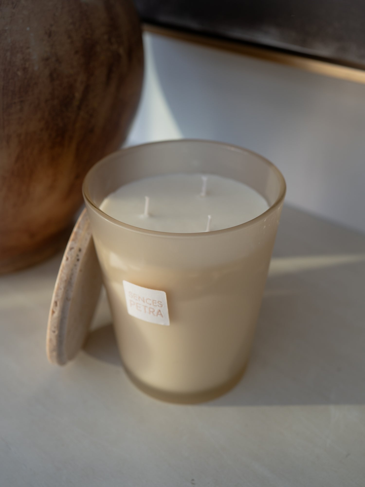 A frosted glass jar candle with cream-colored wax sits on a light surface with a travertine lid balanced against the side, with a large brown ceramic vessel in the background
