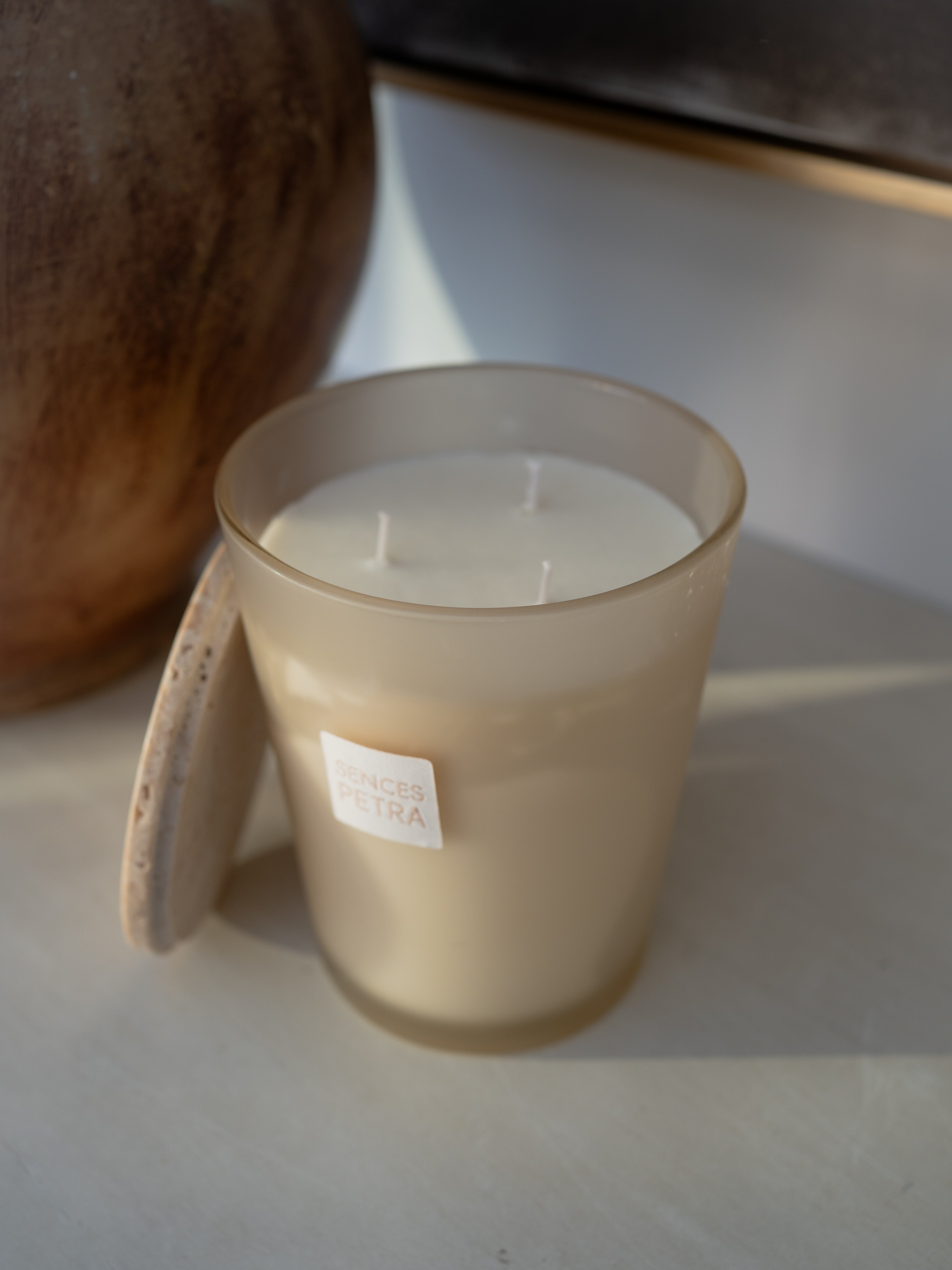 A frosted glass jar candle with cream-colored wax sits on a light surface with a travertine lid balanced against the side, with a large brown ceramic vessel in the background