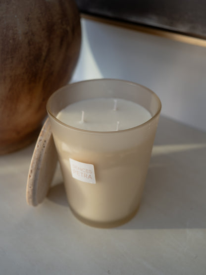 A frosted glass jar candle with cream-colored wax sits on a light surface with a travertine lid balanced against the side, with a large brown ceramic vessel in the background