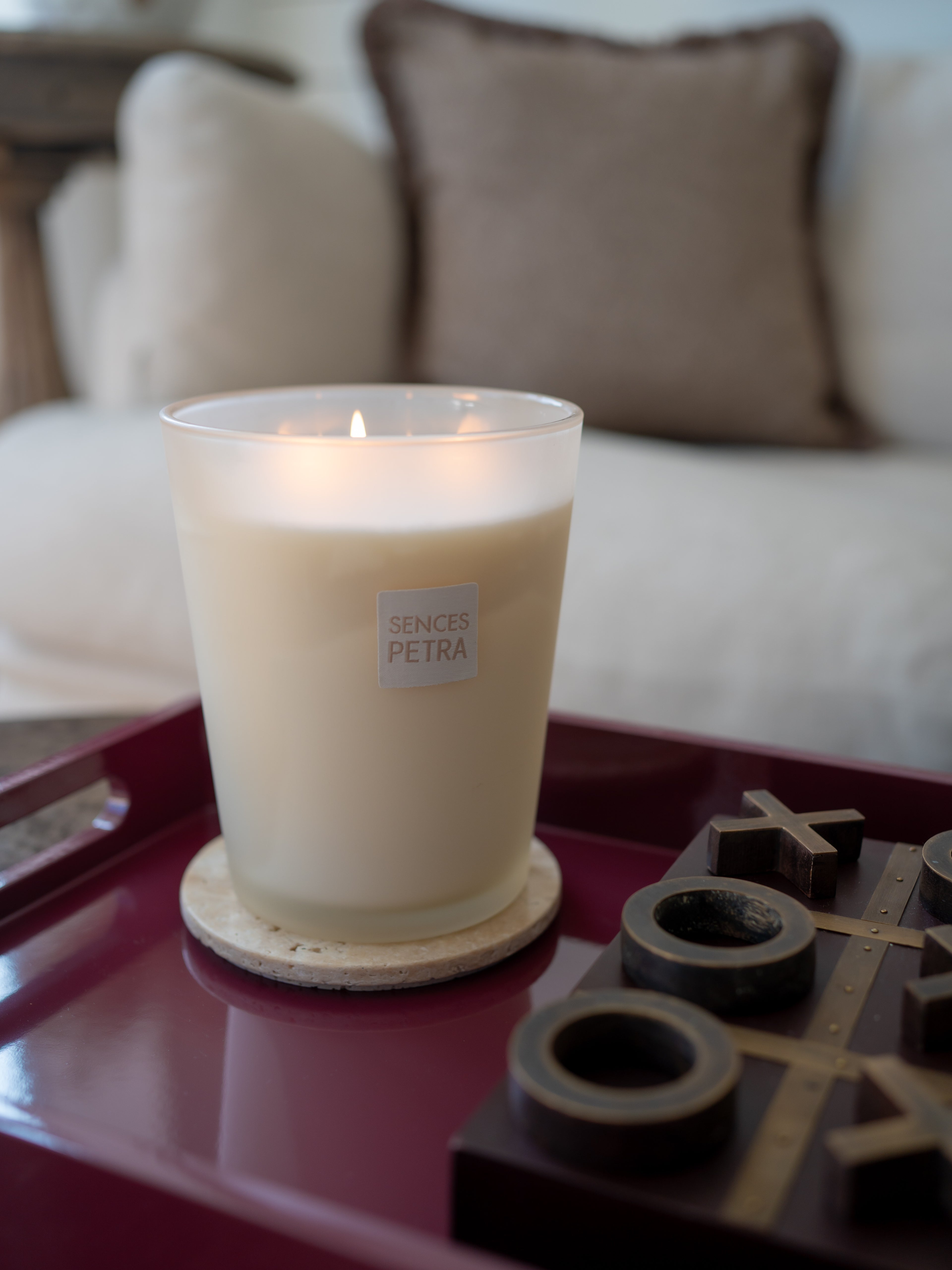 A lit Sences Petra candle in a frosted glass jar sits on a wooden coaster on a dark burgundy tray, with a wooden game board and blurred pillows in the background.