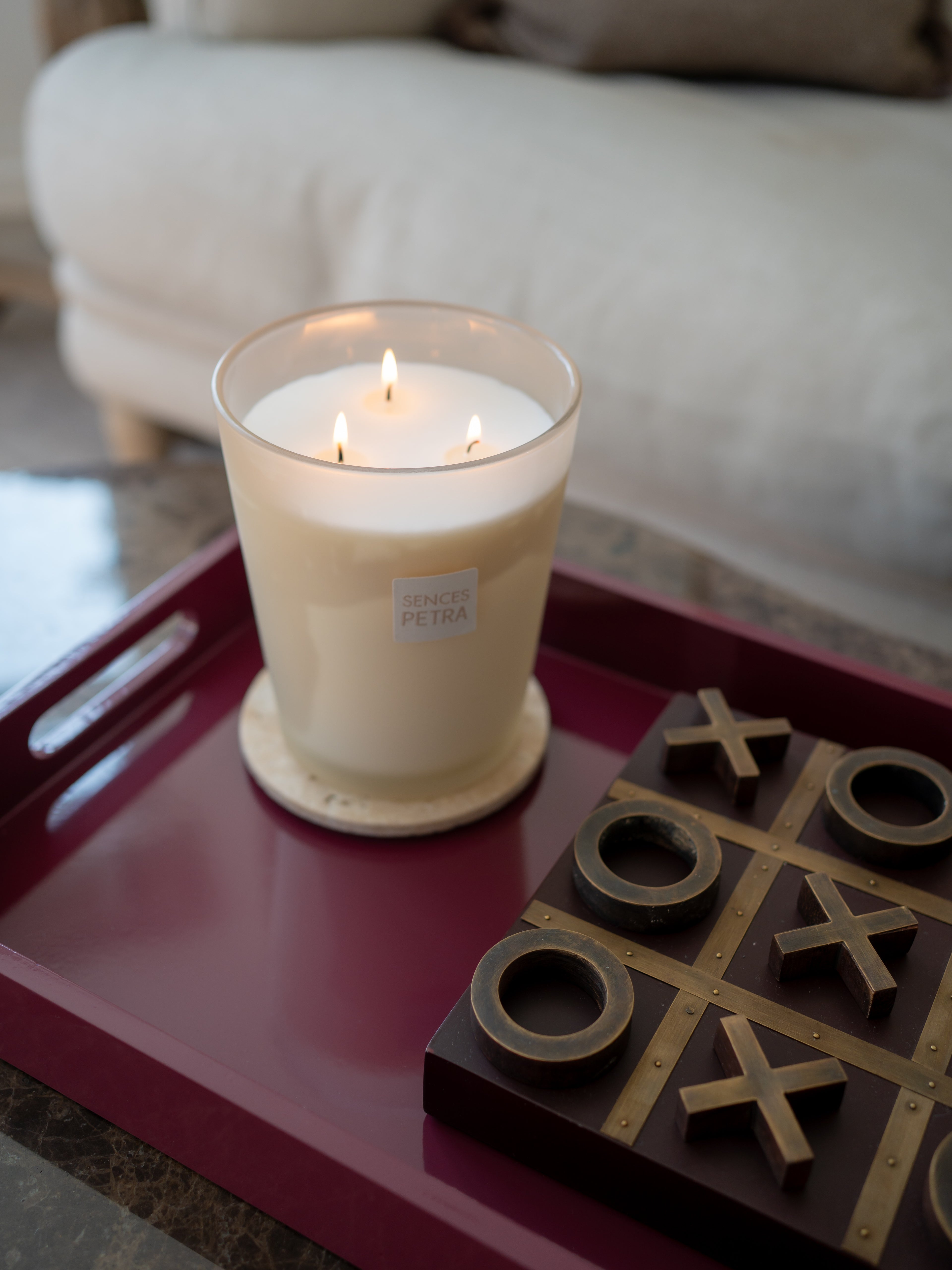A lit white candle in a clear glass jar sits on a dark burgundy tray next to a wooden tic-tac-toe game board with brass-colored pieces. The background shows a softly blurred living room setting with a light-colored sofa.