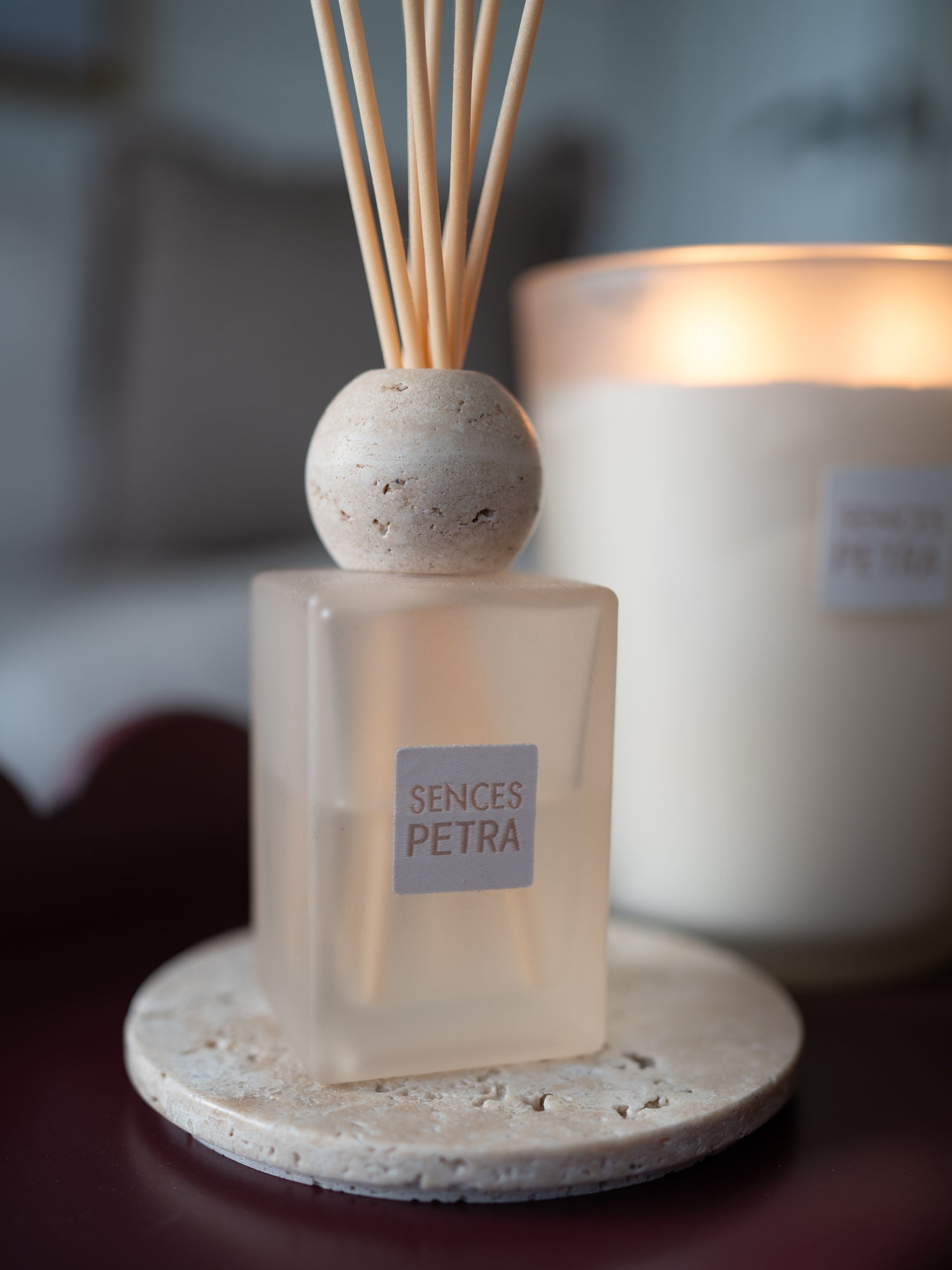Scented reed diffuser bottle with a round stone stopper and natural reeds, placed on a round stone coaster in front of a softly blurred lit candle. The diffuser bottle has a square frosted base with a label reading &