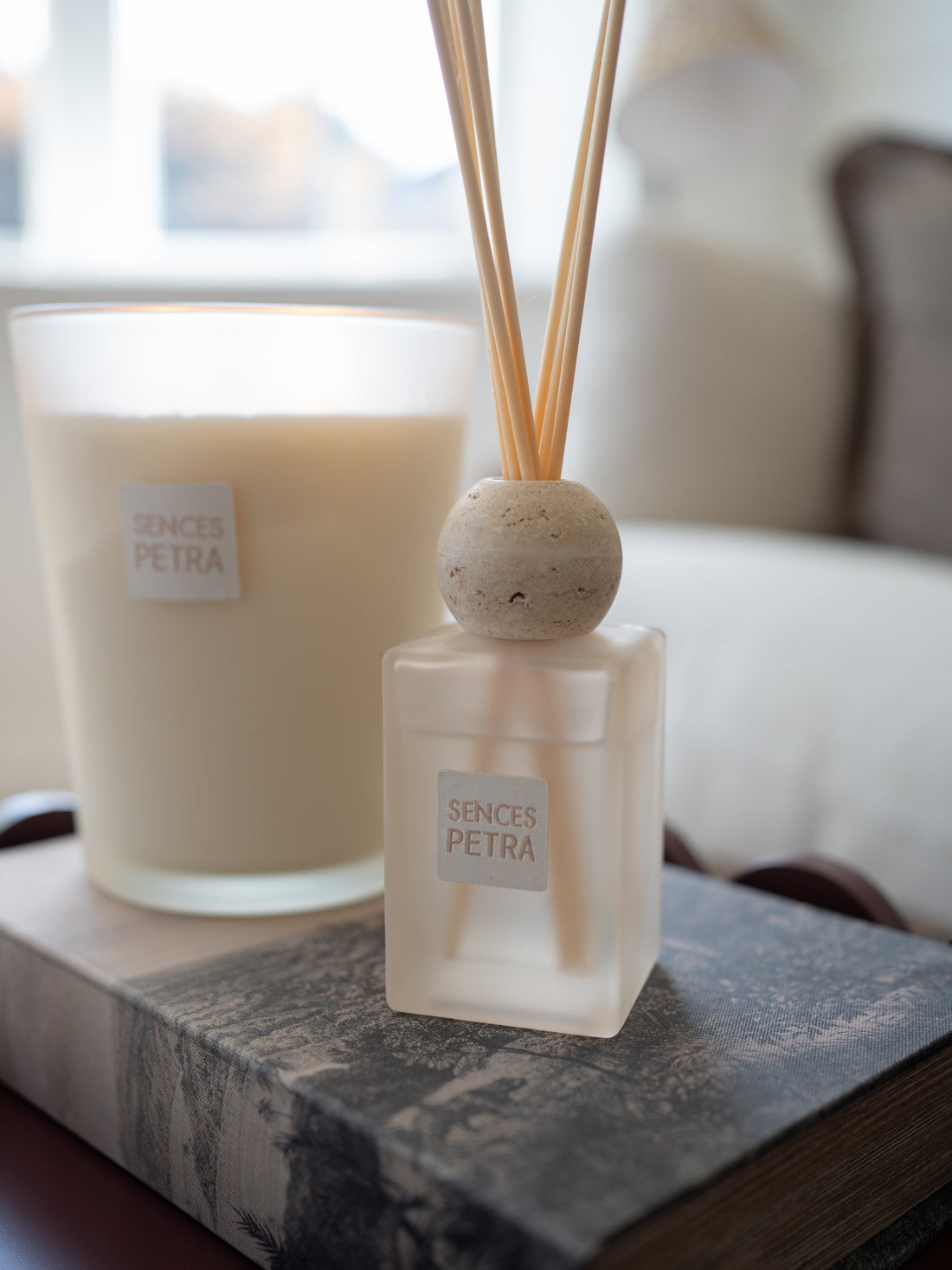 A frosted glass reed diffuser bottle with natural wood reeds and a round stone stopper sits on a dark marble book, with a matching frosted candle in the background.