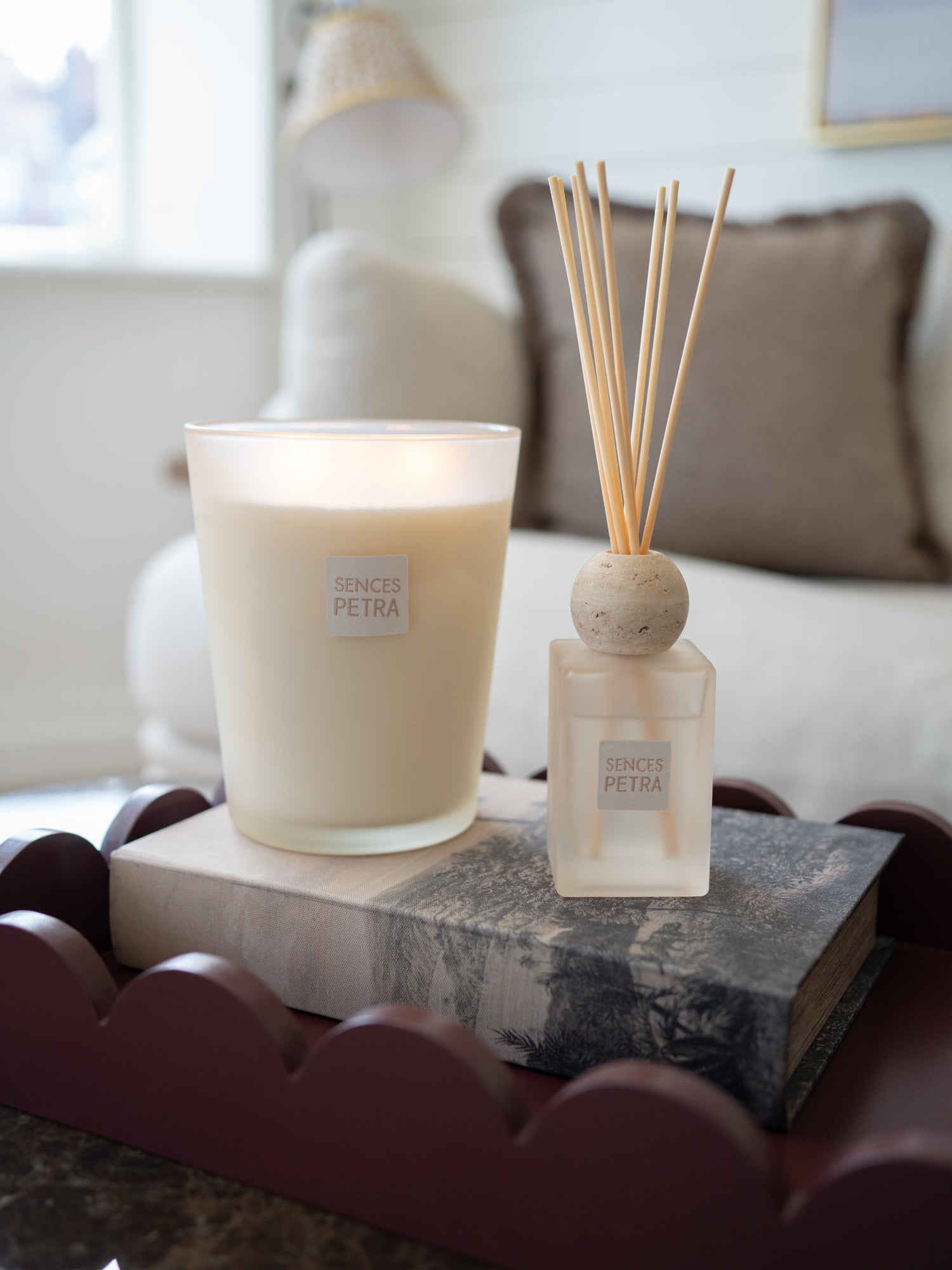 A lit Senses Petra candle in a frosted glass jar sits on a gray book next to a matching Senses Petra reed diffuser with wooden reeds, styled on a dark scalloped tray in a softly blurred living room setting.