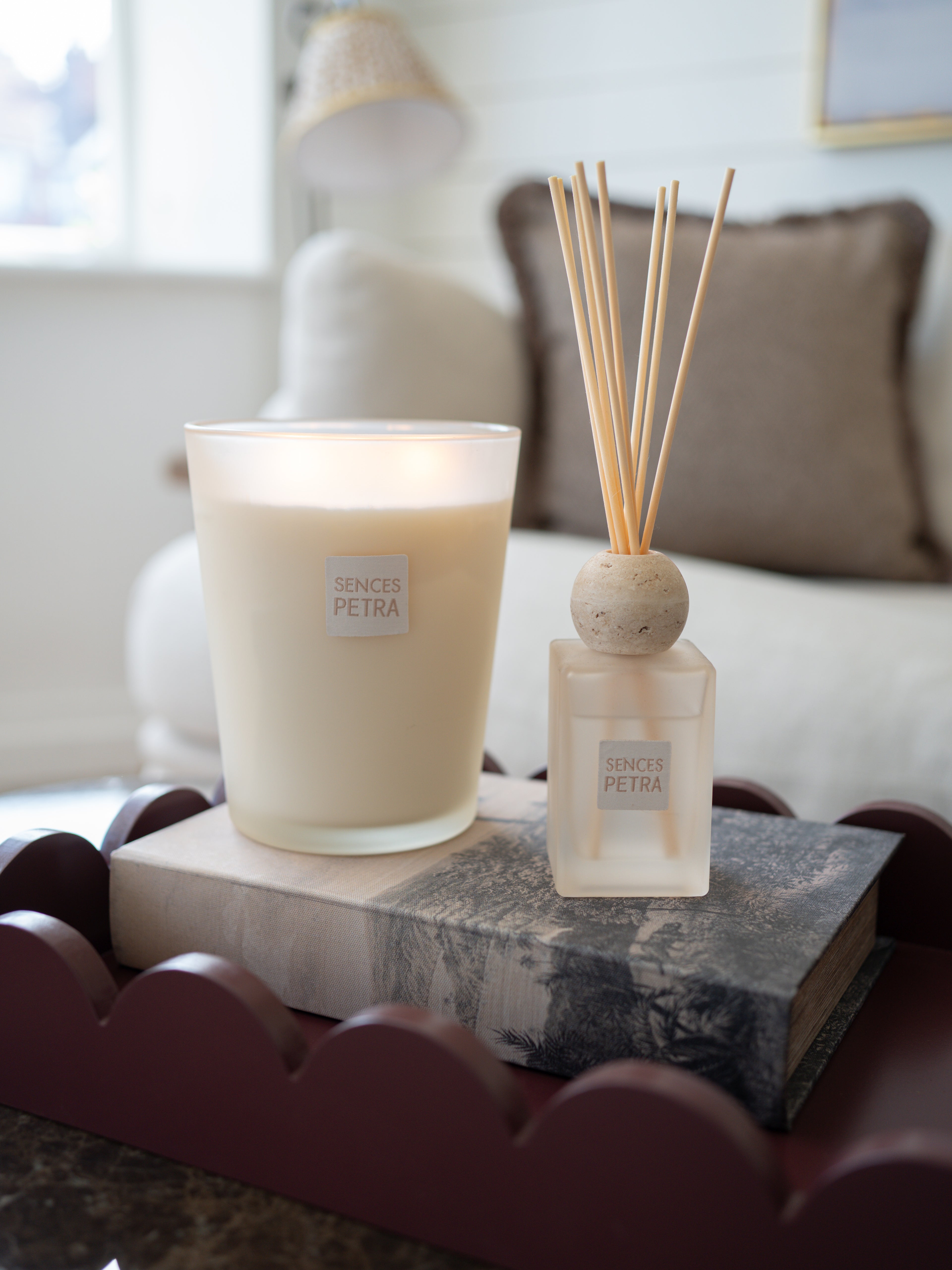 A lit Senses Petra candle in a frosted glass jar sits on a gray book next to a matching Senses Petra reed diffuser with wooden reeds, styled on a dark scalloped tray in a softly blurred living room setting.