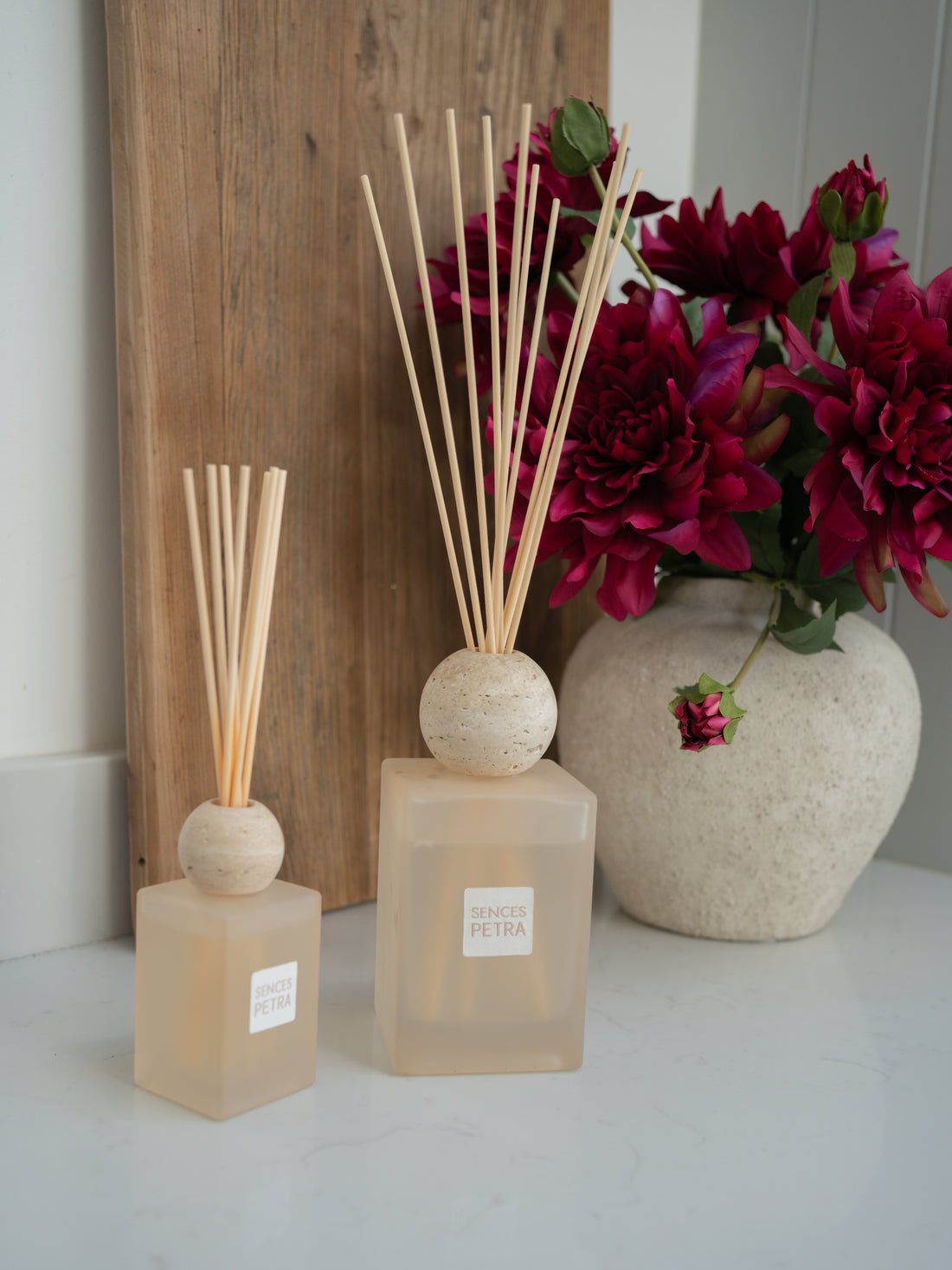 Two luxury reed diffusers with reeds and a vase of flowers on a white surface.