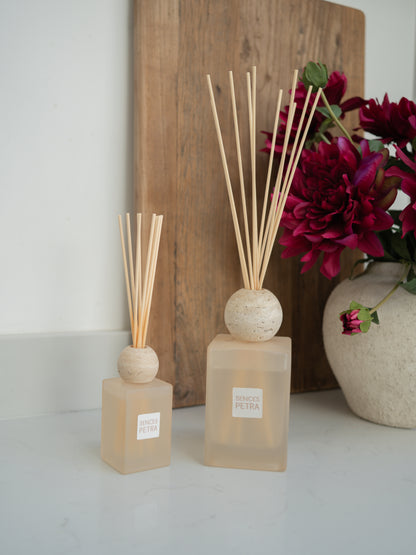 Two diffusers with reeds on a white surface with a wooden board and flowers in the background.