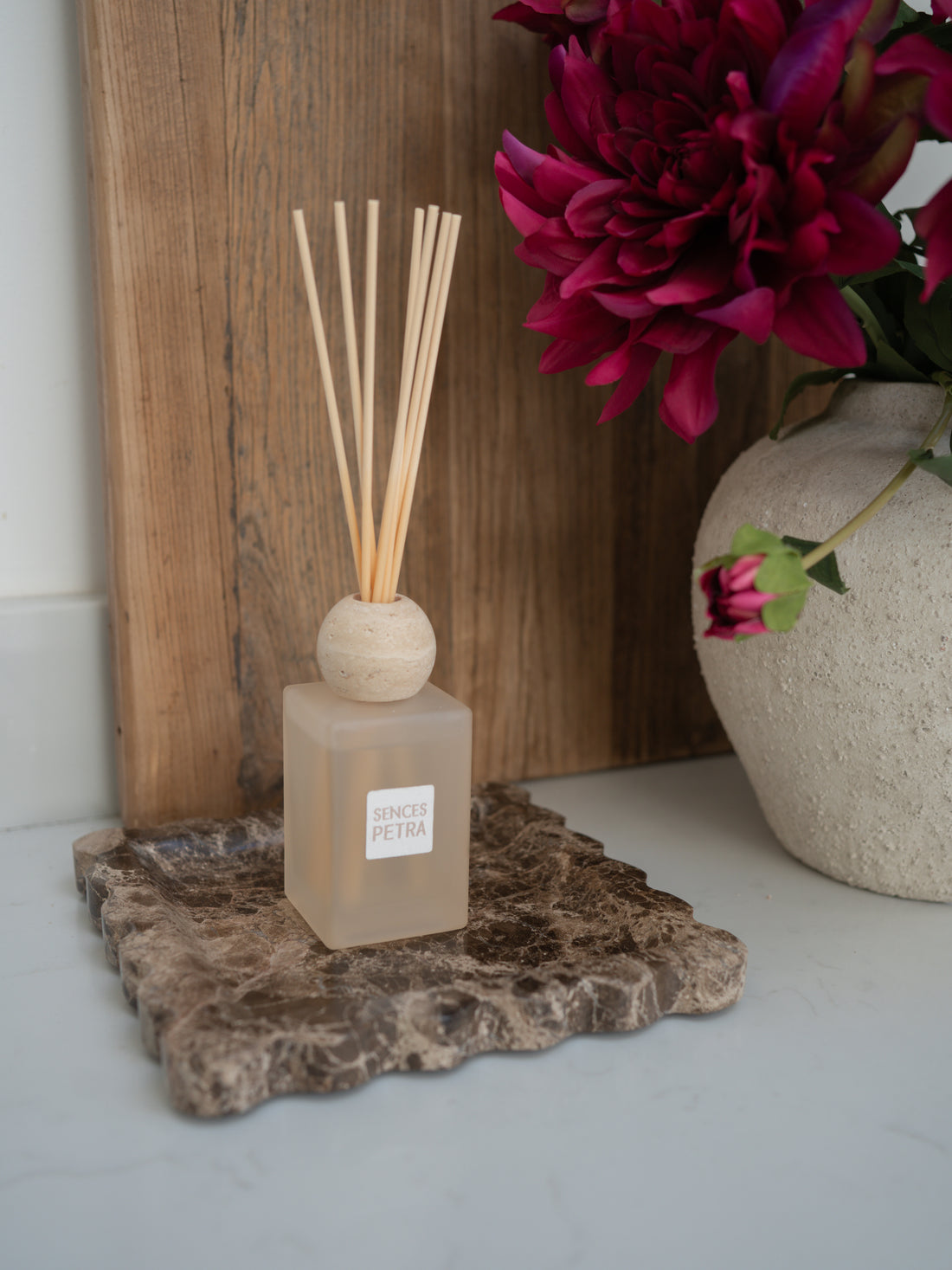 Reed diffuser on a textured brown marble scalloped tray surface with a wooden board and flowers in the background