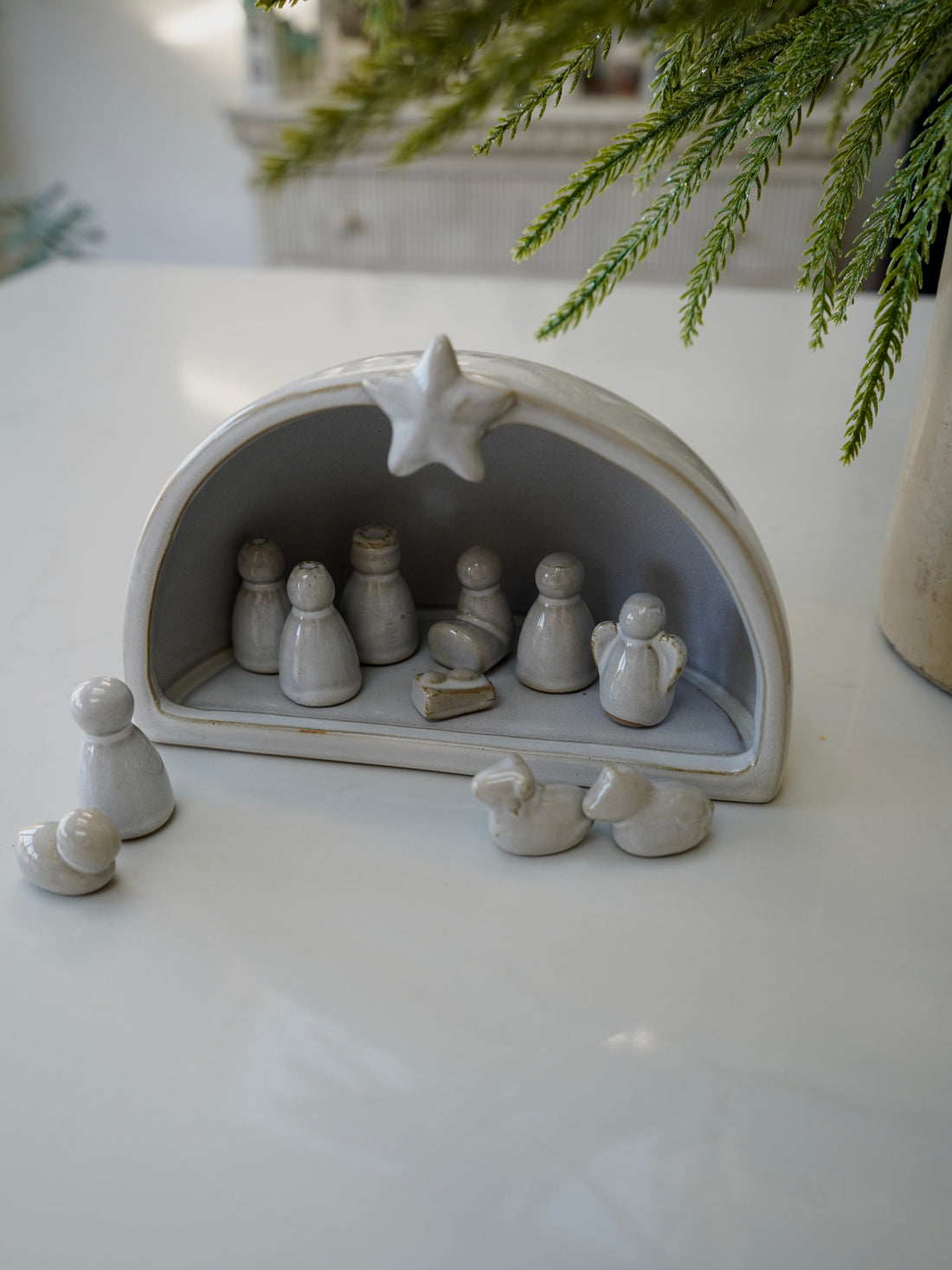 Small nativity scene set in a gray stable on a white surface with greenery in the background.