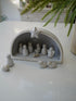 Small nativity scene set in a gray stable on a white surface with greenery in the background.