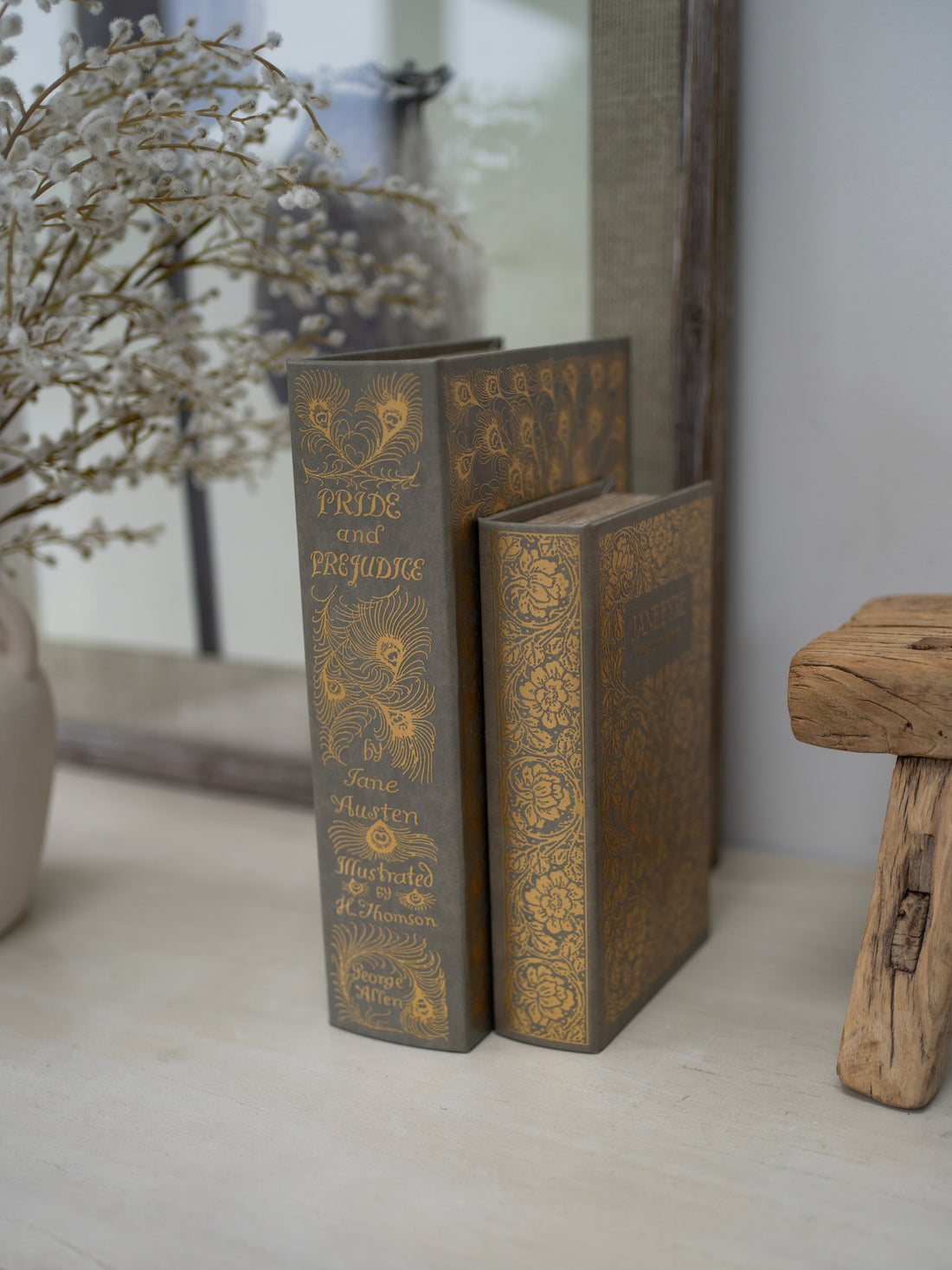 Decorative books with gold embossed text on a surface with a mirror and plant in the background