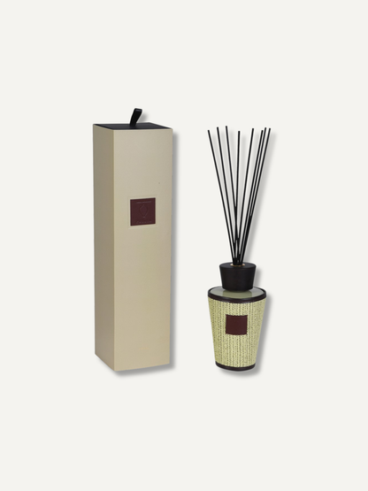 A reed diffuser set shown next to its tall rectangular packaging box. The diffuser features a textured beige glass bottle with a black cap, black reeds, and a small dark brown square label.