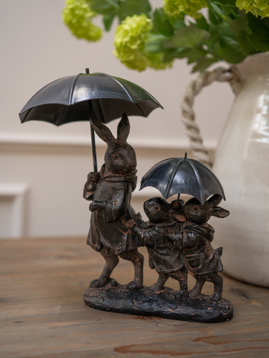 Bronze statue of a rabbit family with an umbrella on a wooden surface with flowers in the background perfect gift idea and home decor ornament