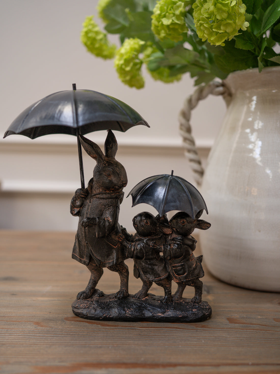 Bronze sculpture of a rabbit with two children under umbrellas, placed on a wooden surface with a vase of flowers in the background beautiful gift idea for animal lovers