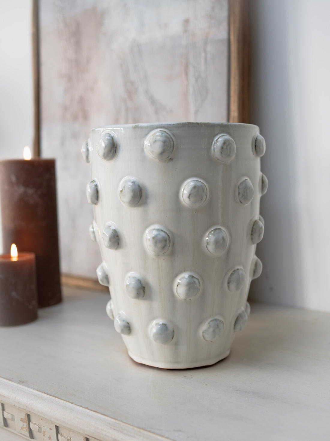 White textured vase on a surface with a candle and framed picture in the background