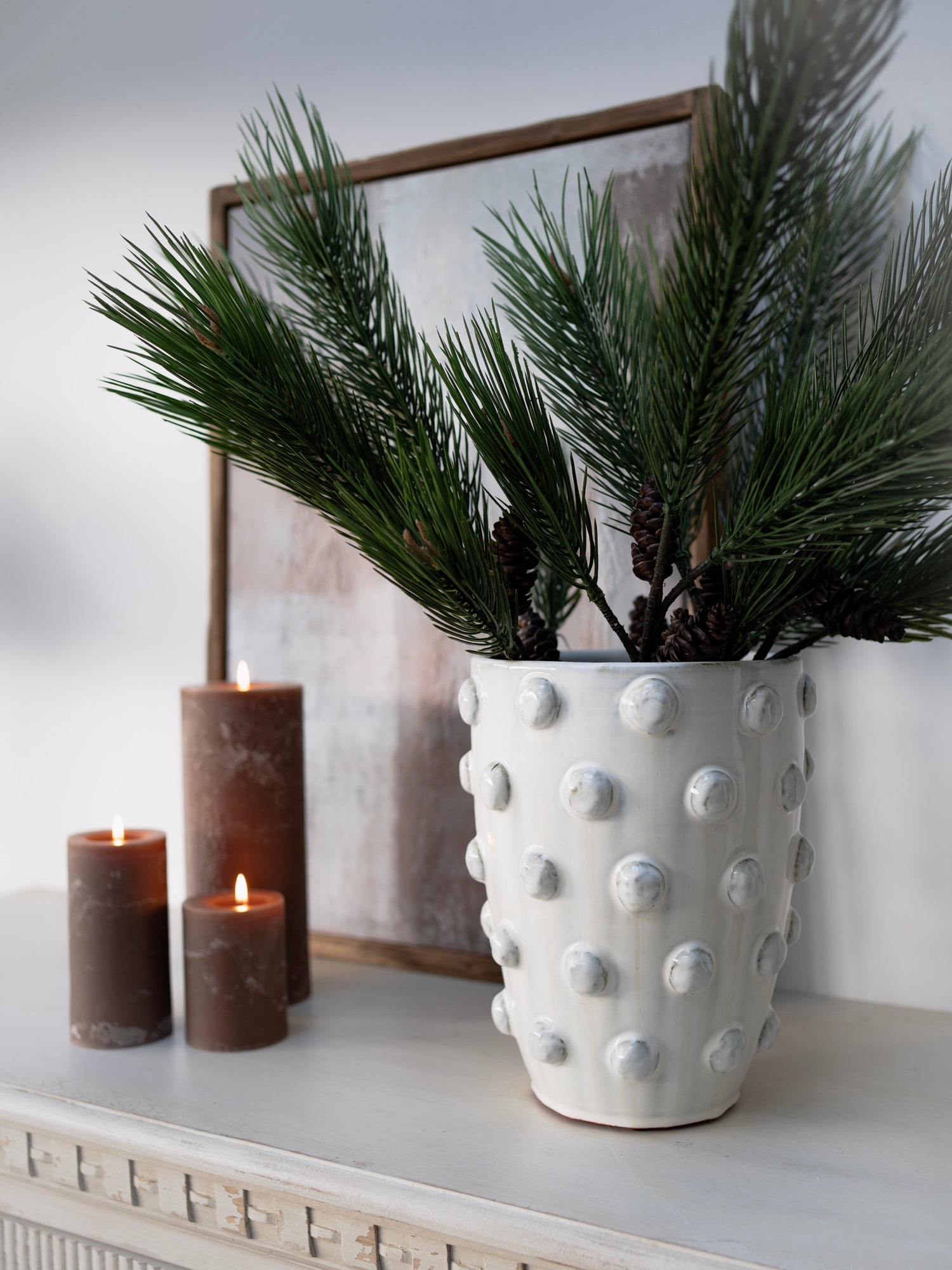 White textured vase with green plant on a shelf with candles and a framed picture in the background