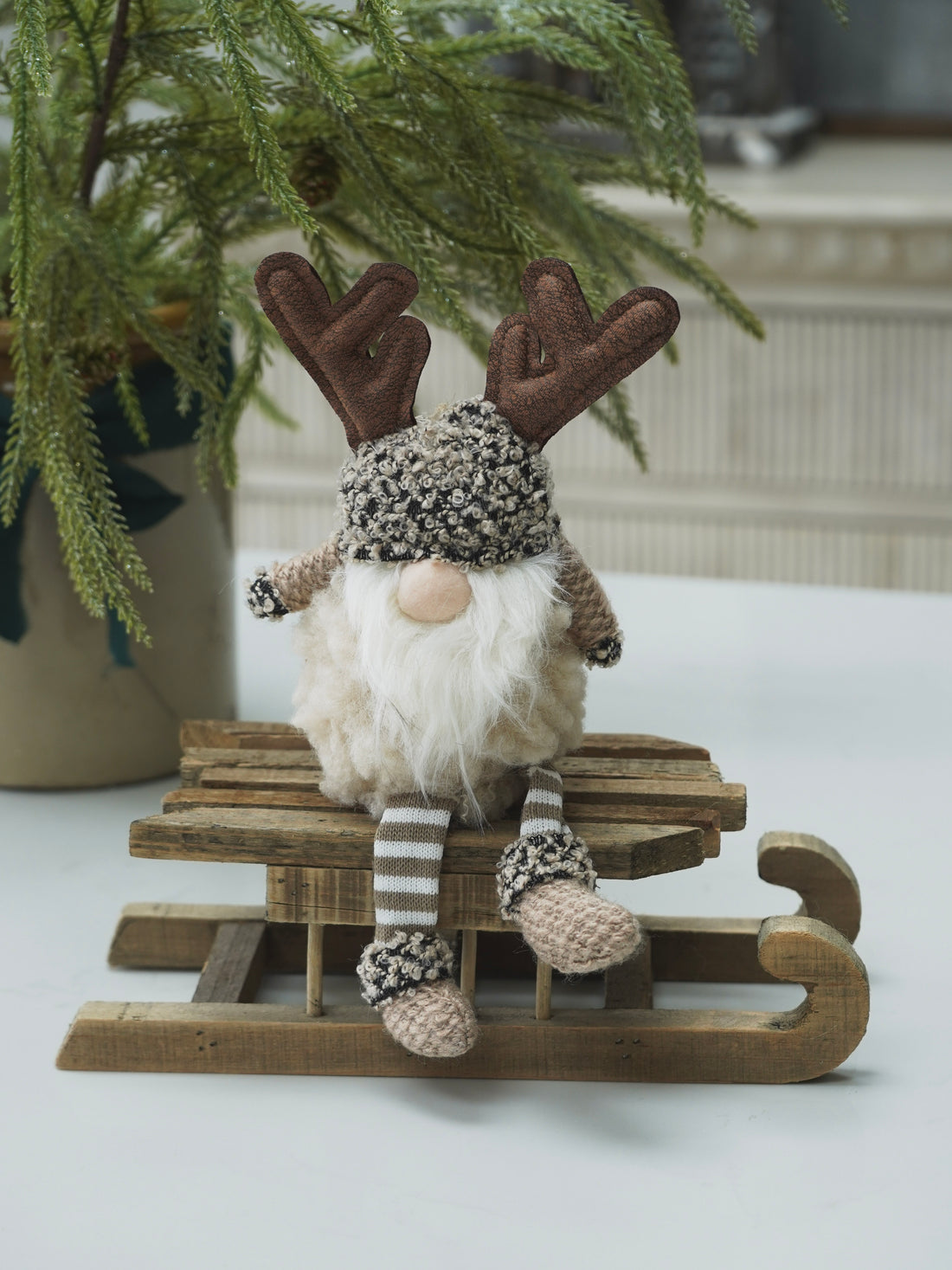 Decorative gnome gonk with reindeer antlers on a small wooden sled, sitting on a white surface with a plant in the background.