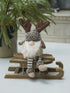 Decorative gnome gonk with reindeer antlers on a small wooden sled, sitting on a white surface with a plant in the background.