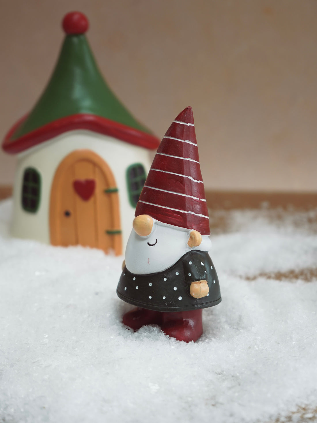 Gnome figurine with a red hat and black outfit standing in front of a small house with a heart door.