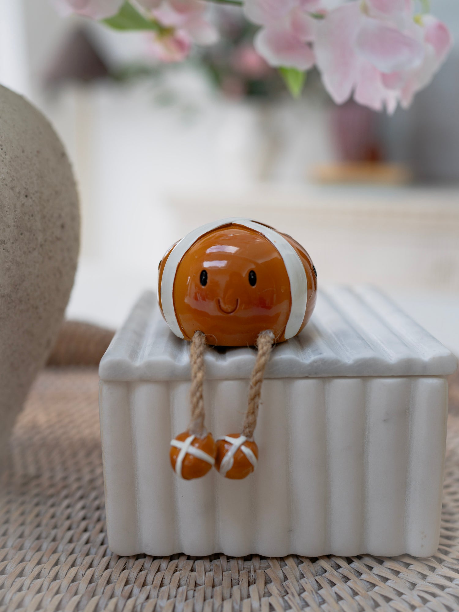 Small ceramic figurine of a hot cross bun on a white box with blurred flowers in the background