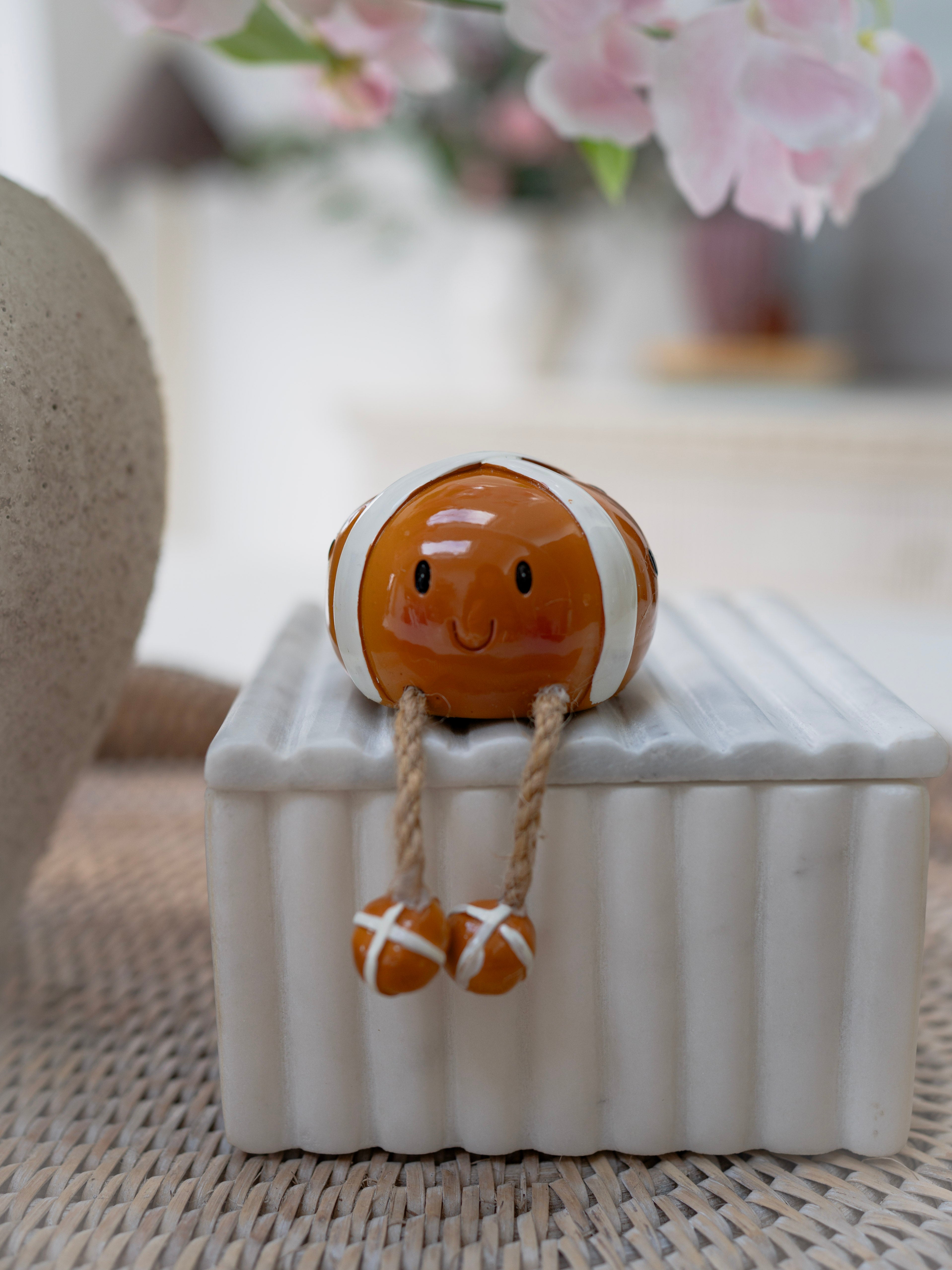 Small ceramic figurine of a hot cross bun on a white box with blurred flowers in the background