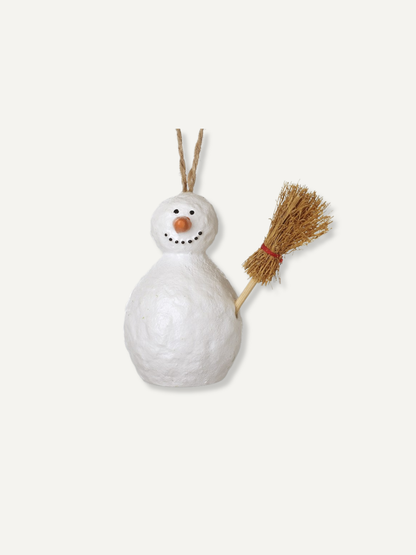 A small white snowman ornament with a textured paper-like finish, a twig nose, black dot eyes and mouth, and a straw-colored broom on its right side. The ornament hangs from two thin twigs against a plain off-white background.