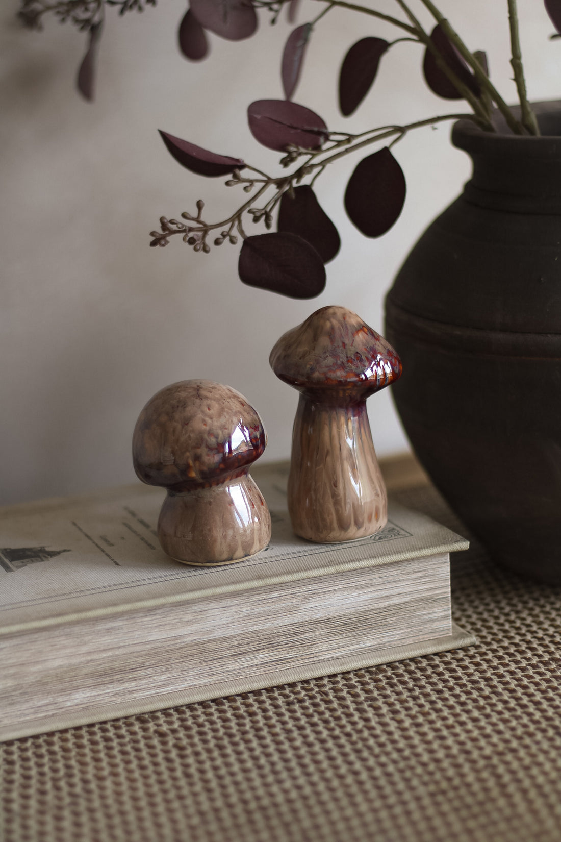 Two ceramic mushroom sculptures on a book with a vase and leaves in the background.