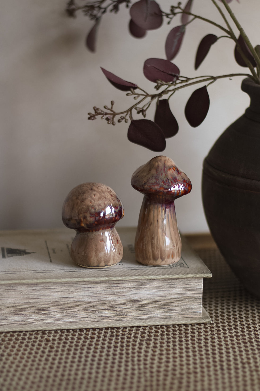 Two ceramic mushroom sculptures on a book with a vase and branches in the background.