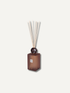 A reed diffuser with a translucent amber-brown glass bottle, a dark round stopper, and natural reed sticks, shown against a solid off-white background.