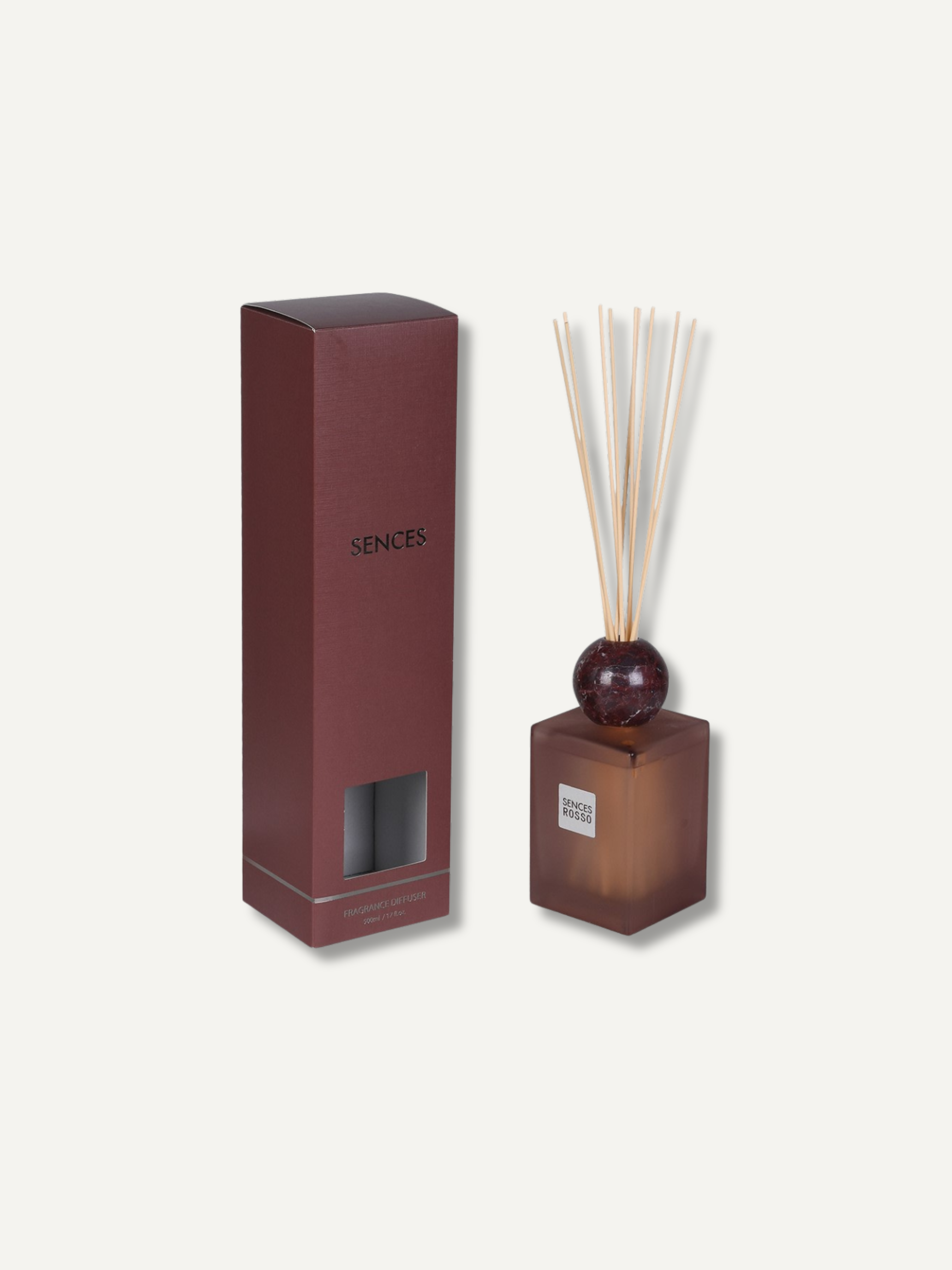 A reed diffuser set with a burgundy glass bottle, natural reeds, and a matching dark brown box, shown on a light beige background. The box is labeled &