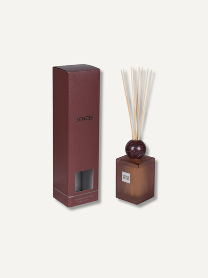 A reed diffuser set with a burgundy glass bottle, natural reeds, and a matching dark brown box, shown on a light beige background. The box is labeled &