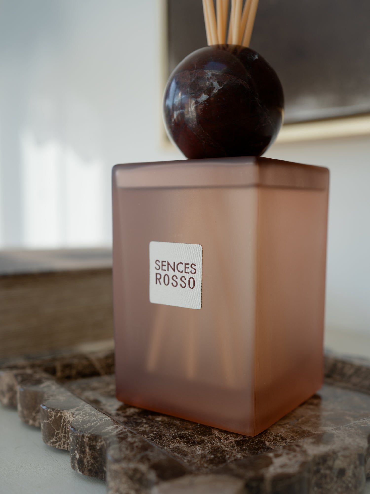 A Sences Rosso reed diffuser with a frosted amber-brown glass bottle, a dark polished stone sphere, and natural reed sticks, displayed on a marble tray in a home setting.