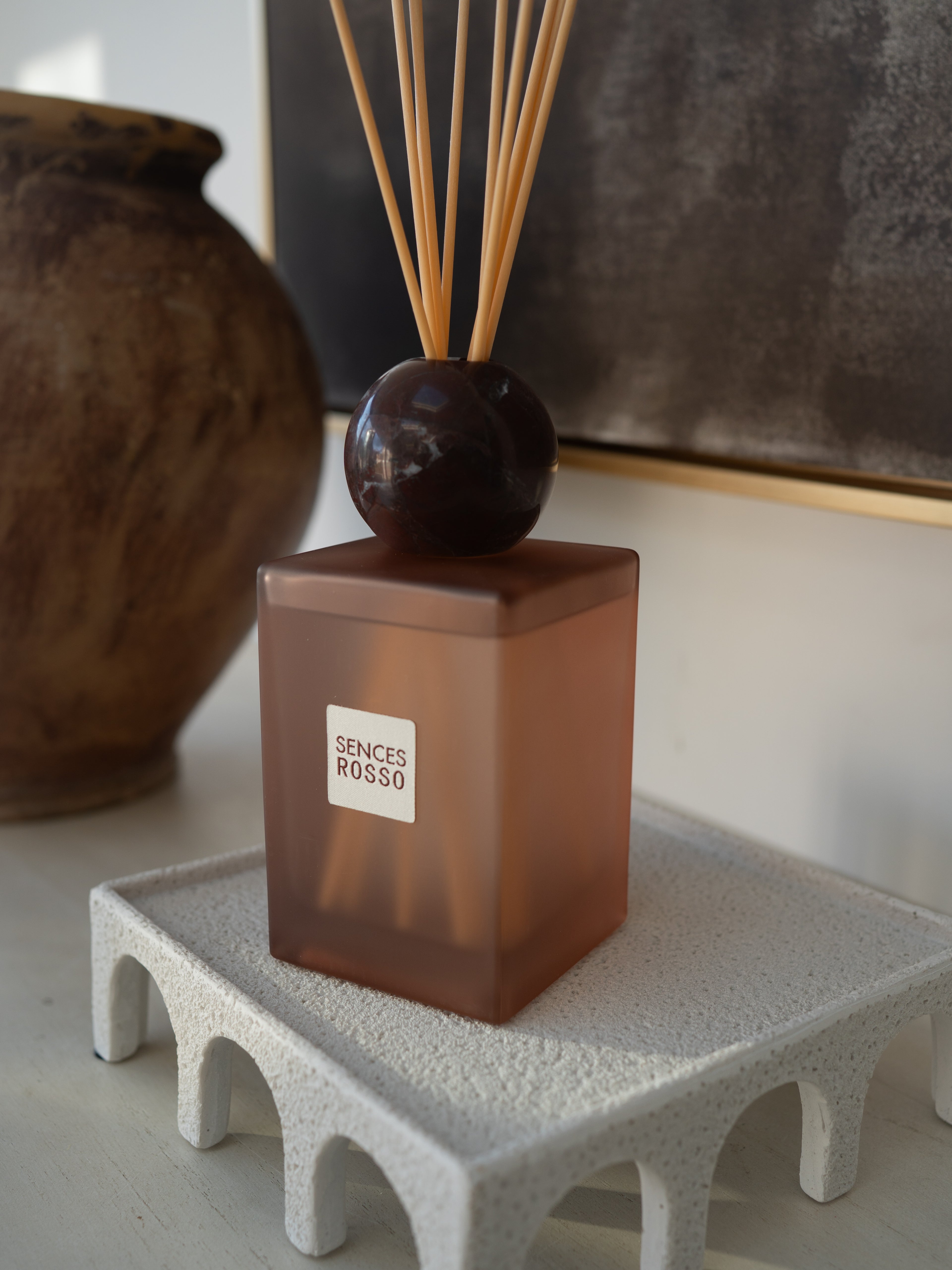A Sences Rosso reed diffuser with a translucent amber-brown cube base, a dark round stopper, and natural reed sticks, displayed on a textured white pedestal in an indoor setting.