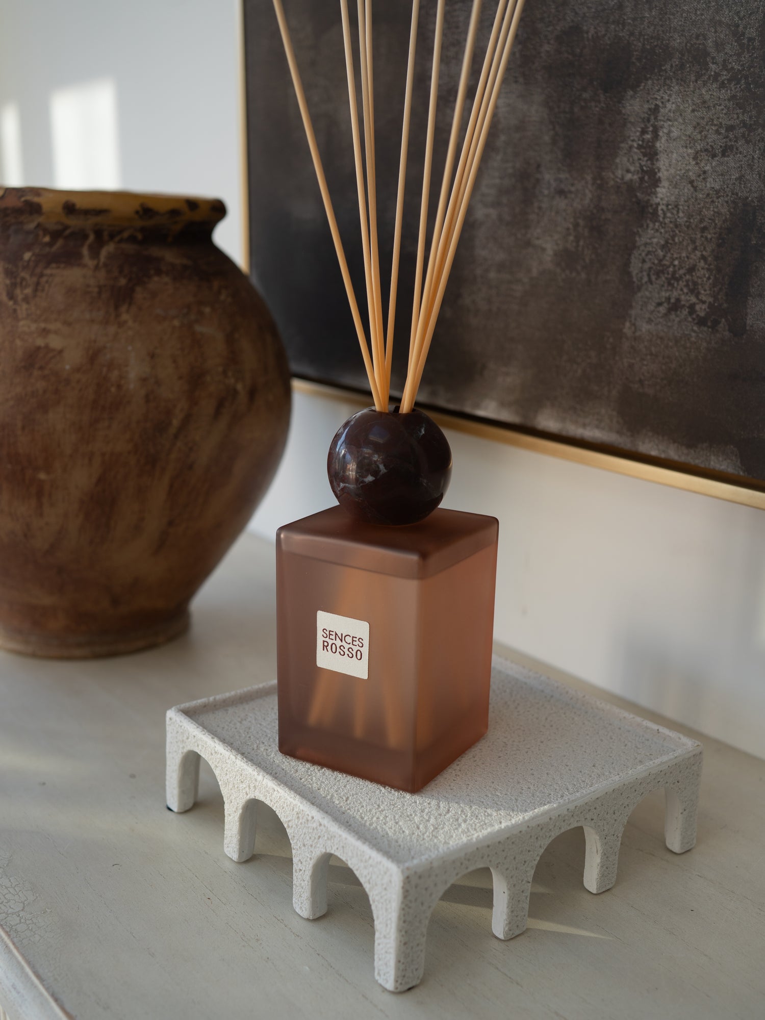 A reed diffuser bottle with a translucent amber-brown rectangular glass body, a dark round marble stopper, and natural reed sticks, displayed on a textured white riser. A large brown ceramic pot and a dark framed artwork are blurred in the background.