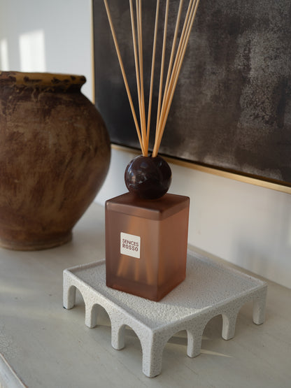 A reed diffuser bottle with a translucent amber-brown rectangular glass body, a dark round marble stopper, and natural reed sticks, displayed on a textured white riser. A large brown ceramic pot and a dark framed artwork are blurred in the background.