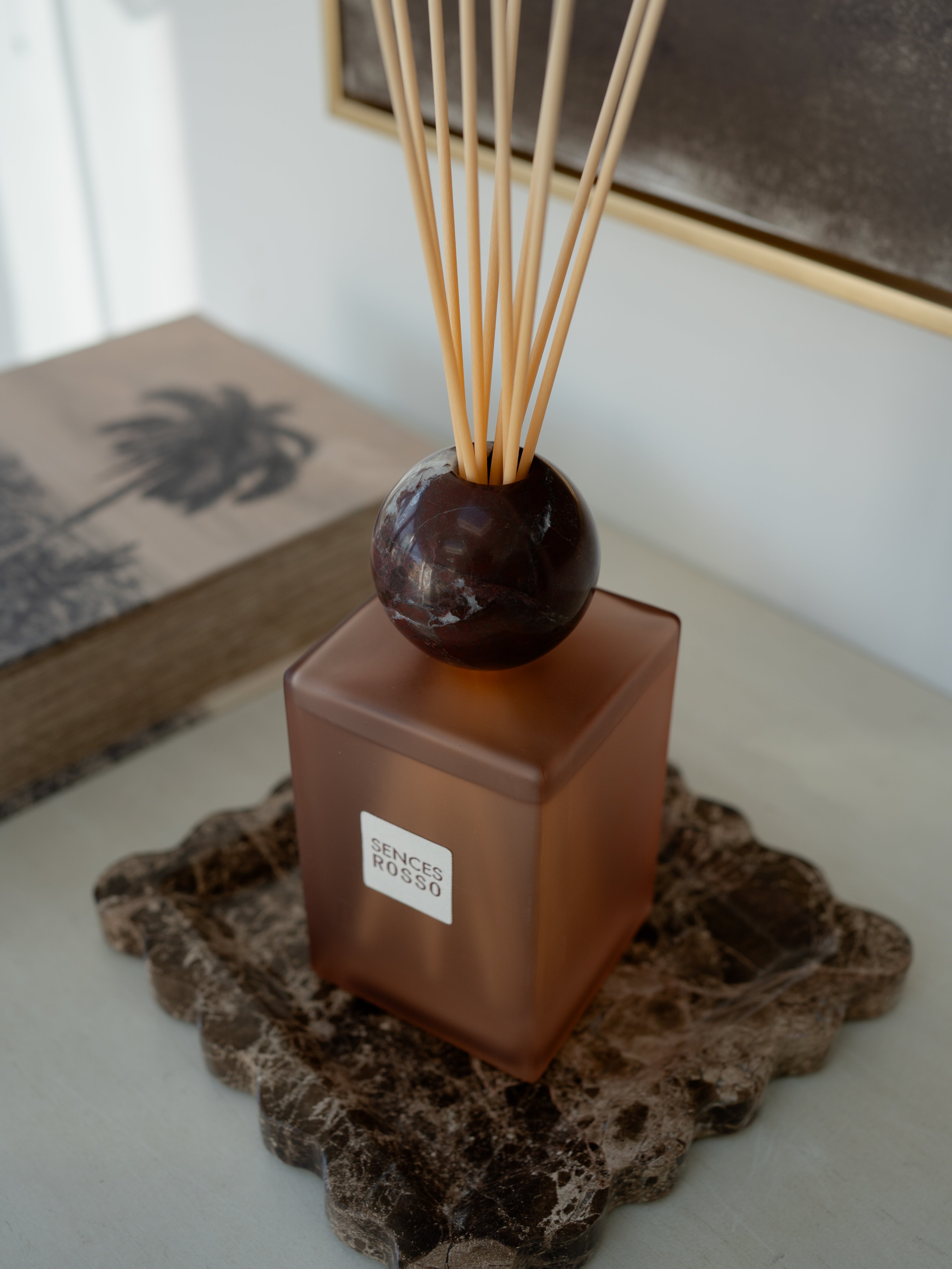 A reed diffuser bottle in frosted amber-brown glass with a dark round stone top and natural reed sticks, displayed on a stone coaster in an indoor setting.