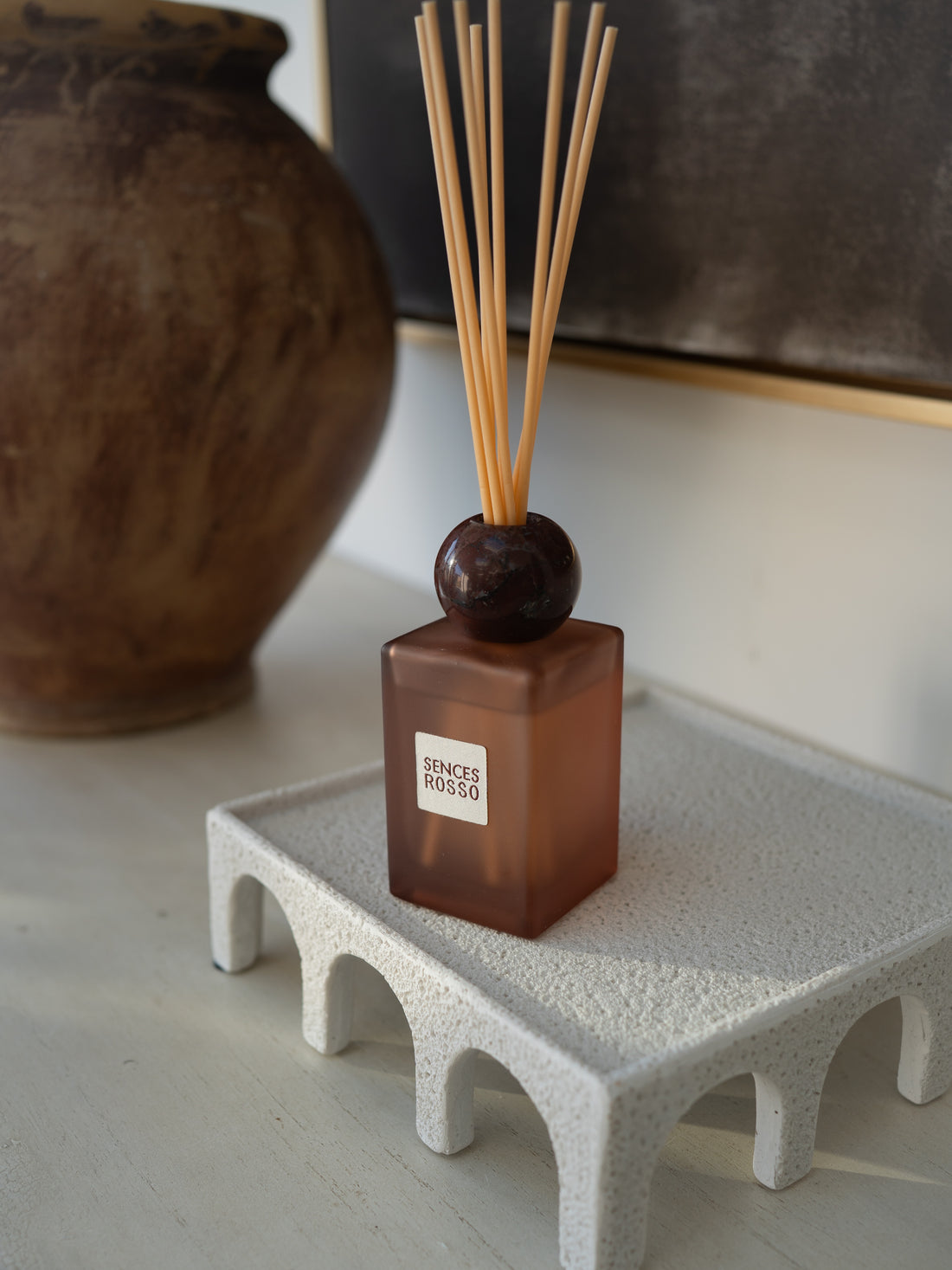 A reed diffuser with a translucent amber-brown glass bottle, dark round stopper, and natural wood reeds, displayed on a textured white tray with a ceramic vase in the background.