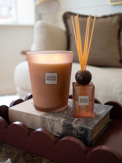 A lit Senses Rosso scented candle in a frosted glass holder sits beside a matching reed diffuser bottle on stacked books in a softly blurred living room setting.