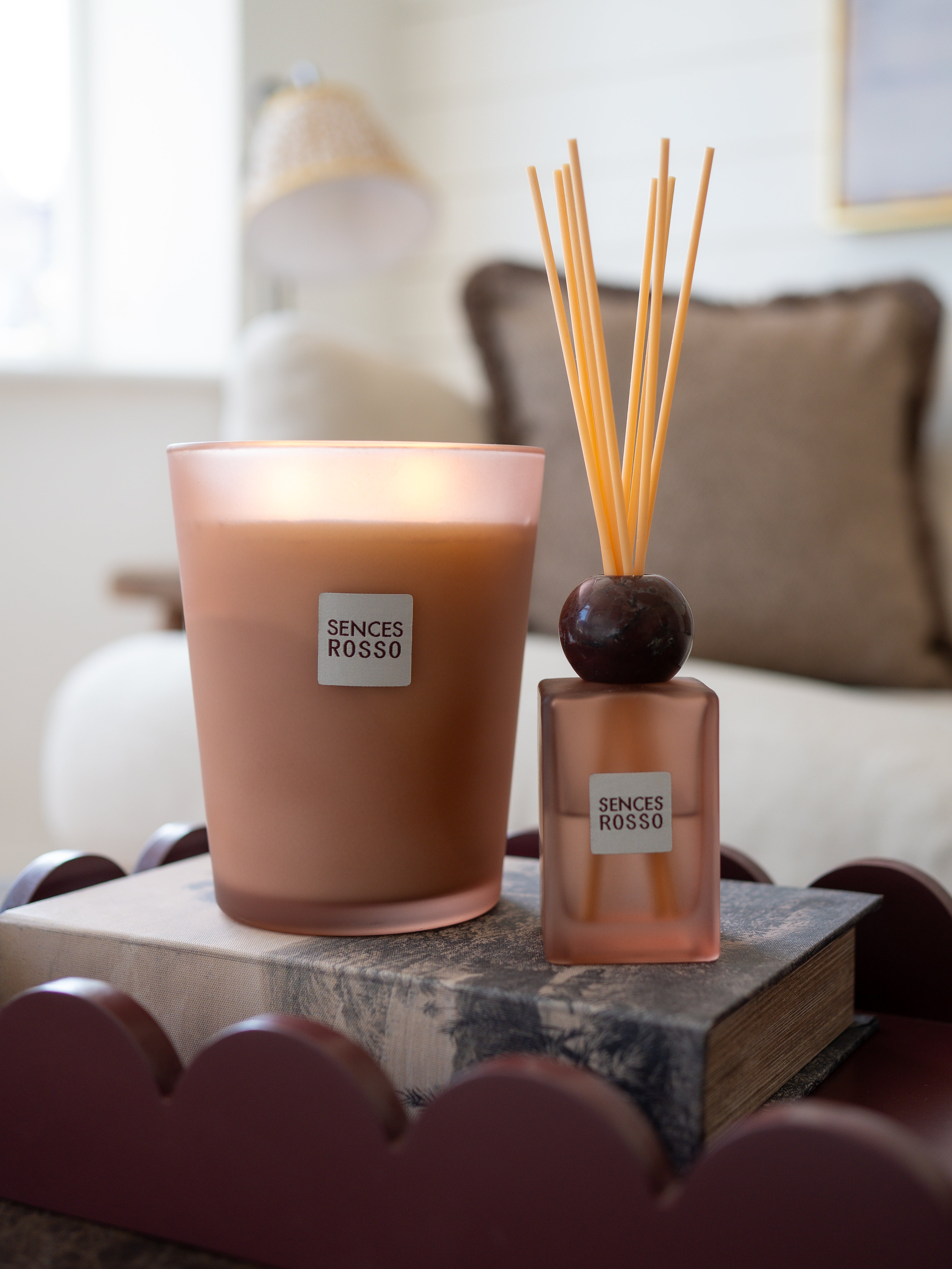 A Sences Rosso scented candle in a frosted glass jar and a matching reed diffuser bottle with wooden reeds, styled on a book in a softly blurred living room setting.